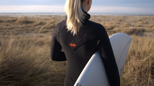 Female surfer wearing the O'Neill Women's Hyperfreak Hooded Chest Zip wetsuit while holding a surfboard and gazing out at the ocean.